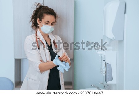 Focused female doctor in protective face mask at medical office putting on rubber gloves preparing for exam of patient at medical office. Young nurse ready to work. Pandemic concept, medical help. 