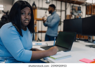 Focused female employee writing in notepad while sitting at desk with laptop in busy open plan office