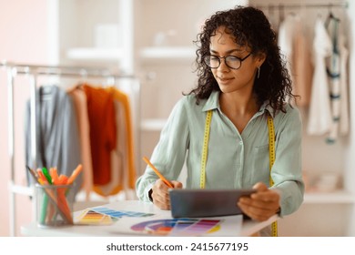 Focused female fashion designer with glasses evaluating color swatches and using a digital tablet in a well-lit modern clothing design studio - Powered by Shutterstock