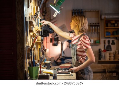 Focused female joiner picking up tool, getting ready for work day. Concentrated unsmiling woman, small carpentry business employee at woodworking creative studio workshop. Artisan professional girl. - Powered by Shutterstock