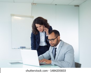 Focused Female Manager Talking To Client On Cellphone. Her Male Colleague Help Her, Using Laptop And Showing Screen To Her. Modern Technology In Business Concept