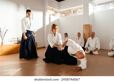 A focused female martial artist practices a wrist lock on a fellow student under the watchful eye of her instructor, as other students observe attentively in a sunlit dojo. - Powered by Shutterstock