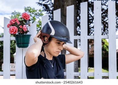 A focused female rider adjusts her helmet before horseback riding, standing by a white fence with potted flowers, preparing for a safe and enjoyable ride. - Powered by Shutterstock