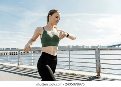Focused female runner checking her smartwatch while training on a sunny waterfront pathway.

