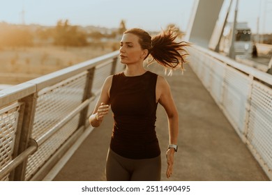 Focused female runner moves gracefully across a bright bridge, enjoying the warmth of the setting sun that casts an inspiring glow on her journey toward fitness - Powered by Shutterstock