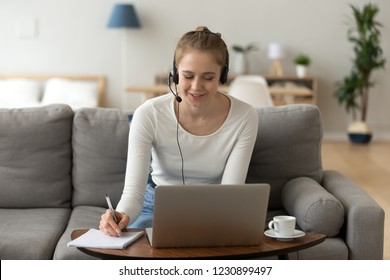 Focused Female Wear Headset Sit On Couch Studying Online At Laptop, Smiling Girl In Earphones Work At Computer At Home, Young Woman Watch Video Seminar Noting Important Data. Distant Education Concept