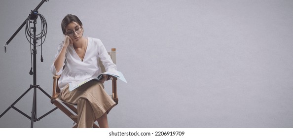 Focused Film Director Sitting On The Director's Chair And Reading A Play Script, Copy Space And White Background