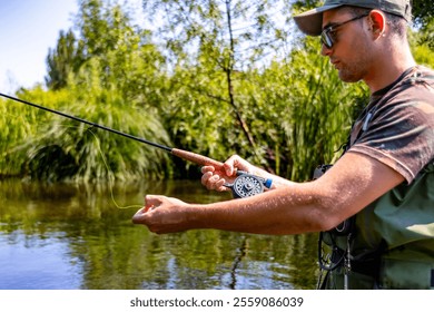 A focused fisherman carefully prepares to cast his line in a calm river, surrounded by lush greenery. The scene captures the quiet concentration of outdoor fishing on a summer day. - Powered by Shutterstock