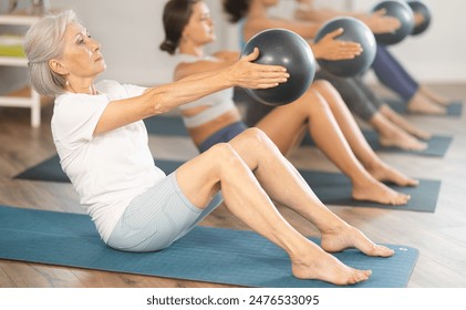 Focused fit senior woman doing sit-ups with stability ball to strengthen abs muscles during group pilates session in spacious sunlit fitness studio.. - Powered by Shutterstock