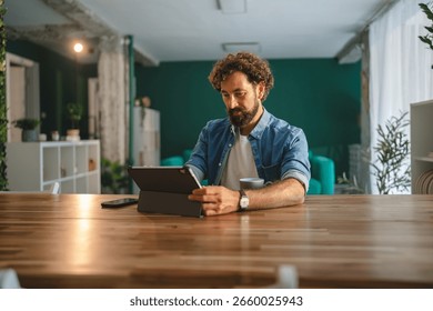 Focused freelancer is using a digital tablet and drinking coffee while working from home in his modern apartment, enjoying the comfort and flexibility of remote work