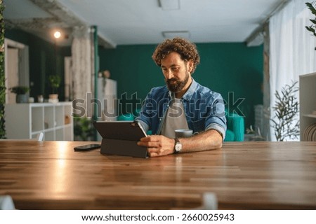 Focused freelancer is using a digital tablet and drinking coffee while working from home in his modern apartment, enjoying the comfort and flexibility of remote work