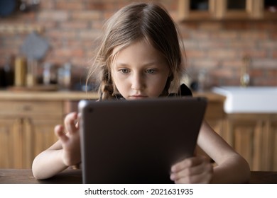 Focused gen Z little schoolkid using online app on tablet, virtual service on computer at home. Serious girl, child with digital device browsing internet, reading electronic book in kitchen