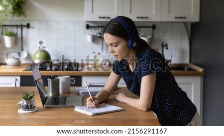 Focused gen Z university student girl in wireless headphones using laptop at kitchen table, writing essay, notes, preparing for test, watching webinar, listening to lecture, attending online lesson