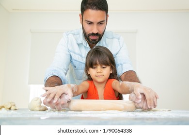 Focused Girl And Her Dad Kneading And Rolling Dough On Kitchen Table With Flour Messy. Father Teaching Daughter To Bake. Front View. Family Cooking Concept