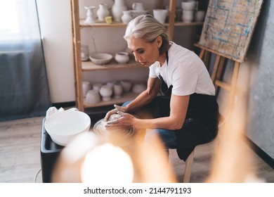 Focused grey hair caucasian female sculptor making clay pot on pottery wheel. Concept of small business and entrepreneurship. Home hobby, entertainment and leisure. Woman working at art studio - Powered by Shutterstock