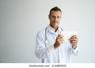 Focused Handsome Doctor Reviewing Patient Test Results. Young Practitioner In White Coat Using Tablet And Posing. Modern Technology In Medicine Concept