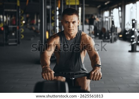 Focused handsome man using rowing equipment in modern gym. Fit young guy exercising on rowing machine at fitness center. Weight loss and cardio fitness concept
