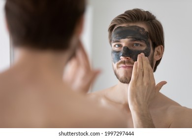 Focused handsome metrosexual guy applying dark cleansing natural clay or mud cosmetic mask on face at mirror for skin treatment, cleaning pores, preventing wrinkles, good complexion. Skincare concept