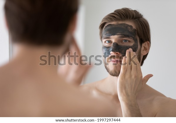 Focused handsome metrosexual guy applying dark\
cleansing natural clay or mud cosmetic mask on face at mirror for\
skin treatment, cleaning pores, preventing wrinkles, good\
complexion. Skincare\
concept