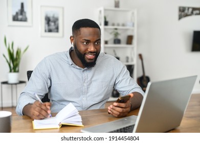 Focused Happy Young African American Man Student Freelancer Making Notes Studying Working With Laptop, Wearing Airpods Looking At The Screen Of The Laptop, Chatting On The Phone At Home Office