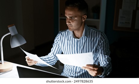 Focused hispanic man reading documents at night in a modern office illuminated by a desk lamp - Powered by Shutterstock