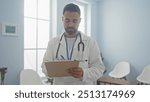 A focused hispanic man wearing a lab coat and stethoscope takes notes in a bright clinic room.