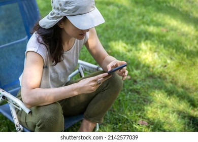 Focused Hispanic Young Woman Wearing A Cap Texting On Mobile Phone And Doing Home Office While Sitting In A Lounge Chair At The Backyard In The Springtime. Copy Space.