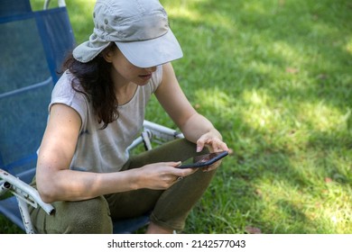 Focused Hispanic Young Woman Wearing A Cap Texting On Mobile Phone And Doing Home Office While Sitting In A Lounge Chair At The Backyard In The Springtime. Copy Space.