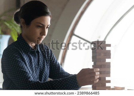 Focused Indian business woman, leader, entrepreneur, startup project manager playing jenga, building carefully shaky tower of wooden blocks, holding stack from falling. Risk, challenge concept