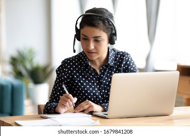 Focused Indian girl wearing headset writing notes in notebook, female student studying language online, listening to lecture, watching trainings, webinar on laptop, sitting at desk - Powered by Shutterstock