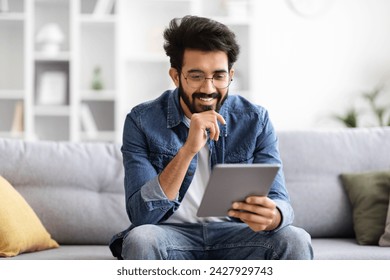 Focused indian man wearing eyeglasses using digital tablet at home, happy smiling eastern male sitting on couch in well-decorated living room, enjoying modern technologies for remote work and leisure - Powered by Shutterstock