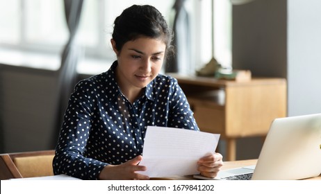 Focused Indian Woman Reading Letter With Important Information, Sitting At Desk With Laptop, Holding Paper Notification With News In Hands, Received Offer Or Invitation, Working With Correspondence