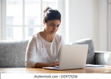 Focused Indian woman using laptop at home, looking at screen, chatting, reading or writing email, sitting on couch, serious female student doing homework, working on research project online