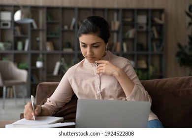 Focused Indian woman write make notes study online on computer in living room. Concentrated serious ethnic female take web training or course on laptop, talk on video call with teacher or tutor.