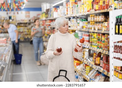 Focused interested gray-haired elderly woman standing near shelves with condiments in supermarket, reading labels on bottles with sauce.. - Powered by Shutterstock