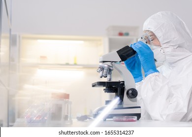 Focused lab technician in protective suit and mask using microscope while working with blood samples