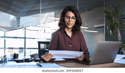 Focused latin hispanic young business woman working on laptop computer reading financial document report in office. Accountant entrepreneur manager businesswoman doing paperwork using pc. Copy space - Powered by Shutterstock