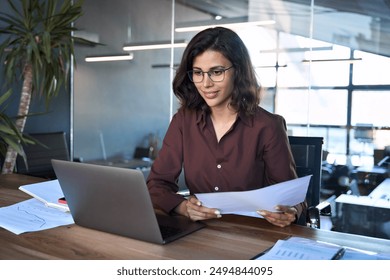 Focused latin hispanic young business woman working on laptop computer reading financial document report in office. Accountant entrepreneur manager businesswoman doing paperwork using pc. Copy space