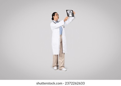 Focused latin woman doctor examining X-ray of the lungs, reflecting a moment of medical evaluation and diagnosis, posing against grey studio background - Powered by Shutterstock