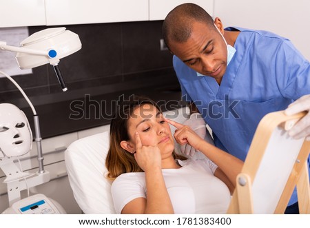 Focused Latina looking at mirror while qualified beautician explaining her future hardware facial procedure in medical esthetic office