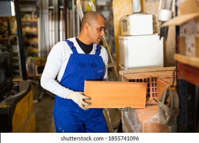 Focused Latino Wearing Blue Coverall Working In Building Materials Hypermarket, Arranging Red Bricks On Display