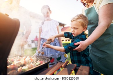 Focused little adorable toddler boy is carefully rotating meat and vegetables on a stick on a grill while being guided by his caring mother and supported by sister. - Powered by Shutterstock