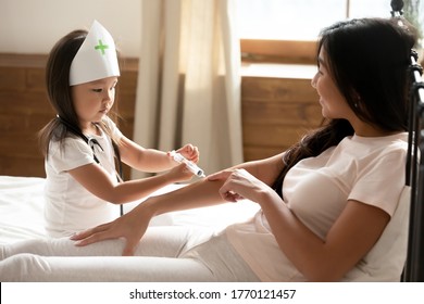 Focused little preschool biracial baby girl in medical cap playing nurse patient with smiling beautiful asian ethnic mother, pretending making prick injection, having fun together in bedroom. - Powered by Shutterstock