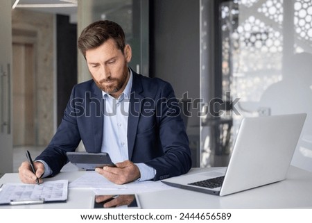 A focused male accountant analyzing paperwork while calculating expenses using laptop and calculator at his office desk.