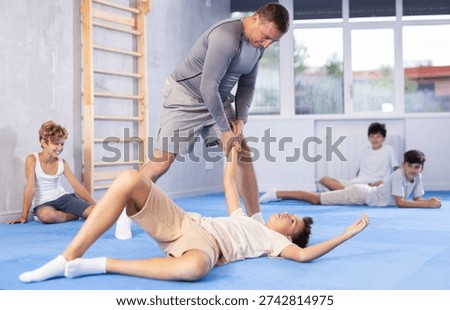 Focused male coach demonstrating basic throws in training bout with teenager to group of interested boys sitting on mat in self-defense training room..