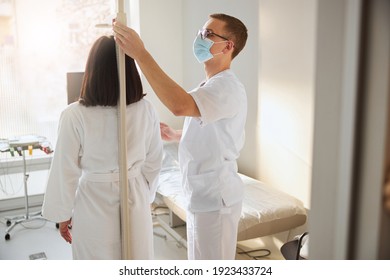 Focused male doctor in a face mask measuring the young woman height at his office