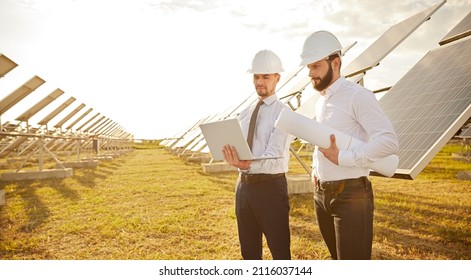 Focused Male Engineers In Hardhats Using Laptop And Discussing Project While Standing Near Photovoltaic Batteries In Countryside