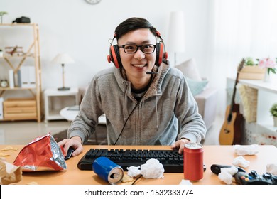 Focused Male Gamer Smiling Looking At Game And Using Computer Keyboard On Messy Desk With Trash. Concentrated Young Asian Man In Headphones Plays Online Home Games On Pc Desktop And Face Camera.