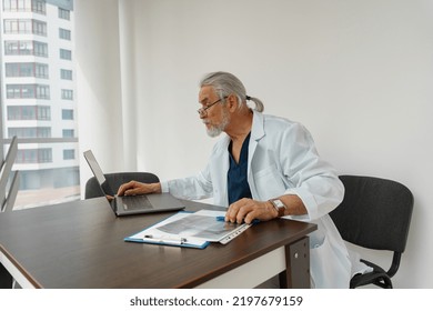 Focused Male Healthcare Worker Doing Paperwork And Using Laptop While Working At Doctor's Office