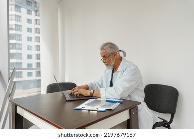 Focused Male Healthcare Worker Doing Paperwork And Using Laptop While Working At Doctor's Office